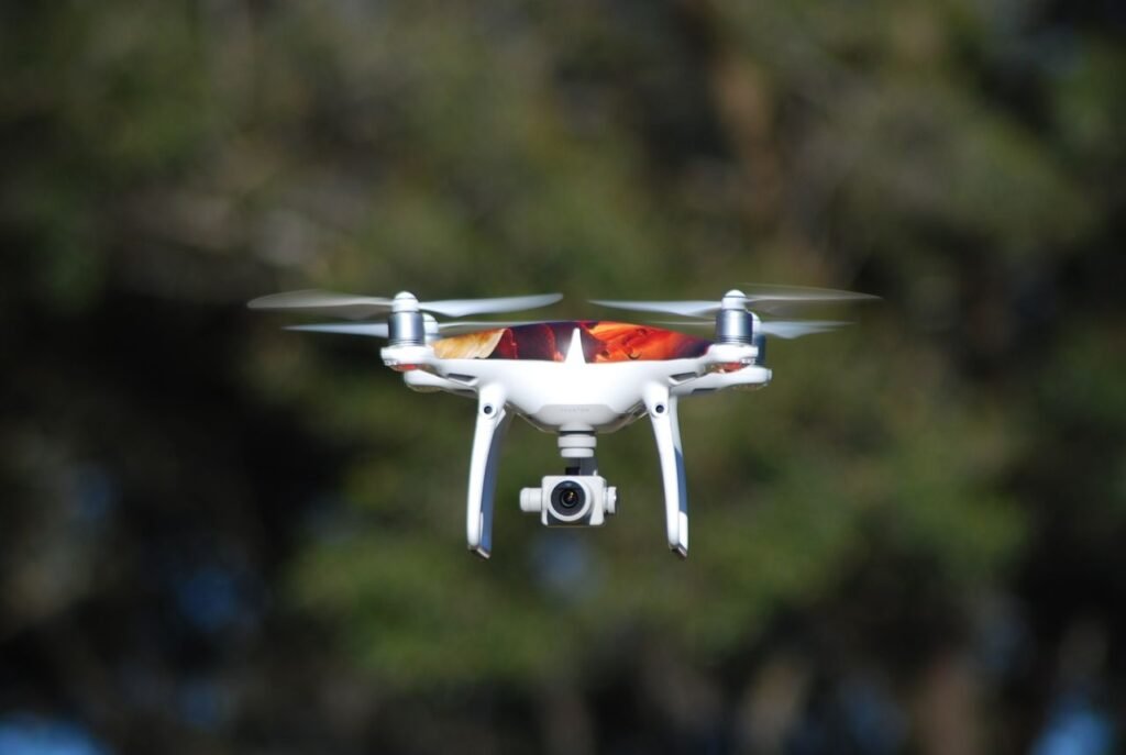 A white drone with camera hovers midair against a blurred natural background.